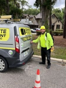 A certified dryer vent technician in a yellow safety helmet standing next to a branded Complete Dryer Vent Services van in front of a residential home.