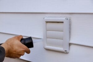A close-up of a person's hand using a black caulking gun to apply white silicone sealant around the edges of a white plastic louvered exhaust vent on white vinyl siding.