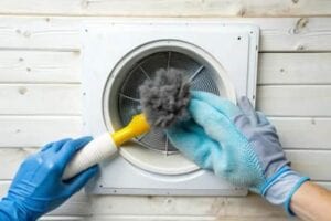 A close-up of a person wearing blue protective gloves using a yellow specialized brush to pull a large clump of grey lint from a circular white dryer vent mounted on a wood-slat wall.