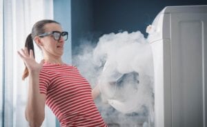 A woman with glasses looking shocked and recoiling as thick white smoke billows out of an open front-loading washing machine in a laundry room.