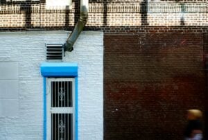 A building exterior wall divided vertically between white textured stone and dark red brick, featuring a blue-framed security door, an industrial exhaust fan, and a metal downspout.
