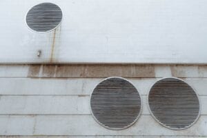 A white tiled and concrete exterior wall featuring three large circular metal ventilation grilles with horizontal slats, showing minor weathering and rust streaks.