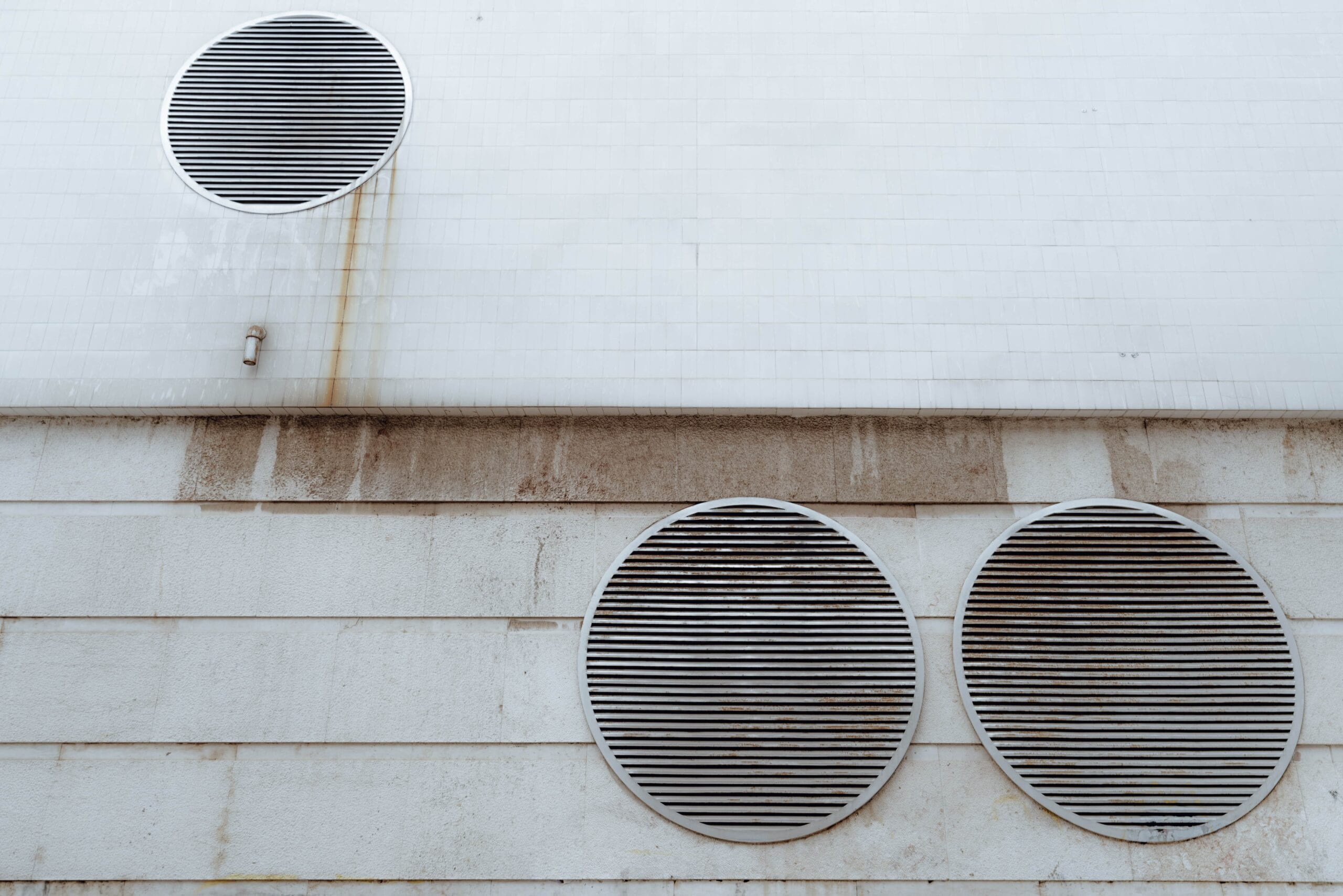 A white tiled and concrete exterior wall featuring three large circular metal ventilation grilles with horizontal slats, showing minor weathering and rust streaks.