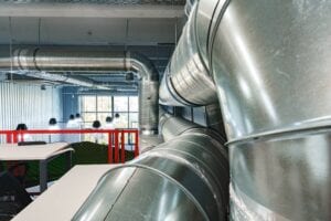 A perspective view of large, shiny silver galvanized steel ventilation ducts and spiral pipes hanging from the ceiling of an industrial or modern office space.