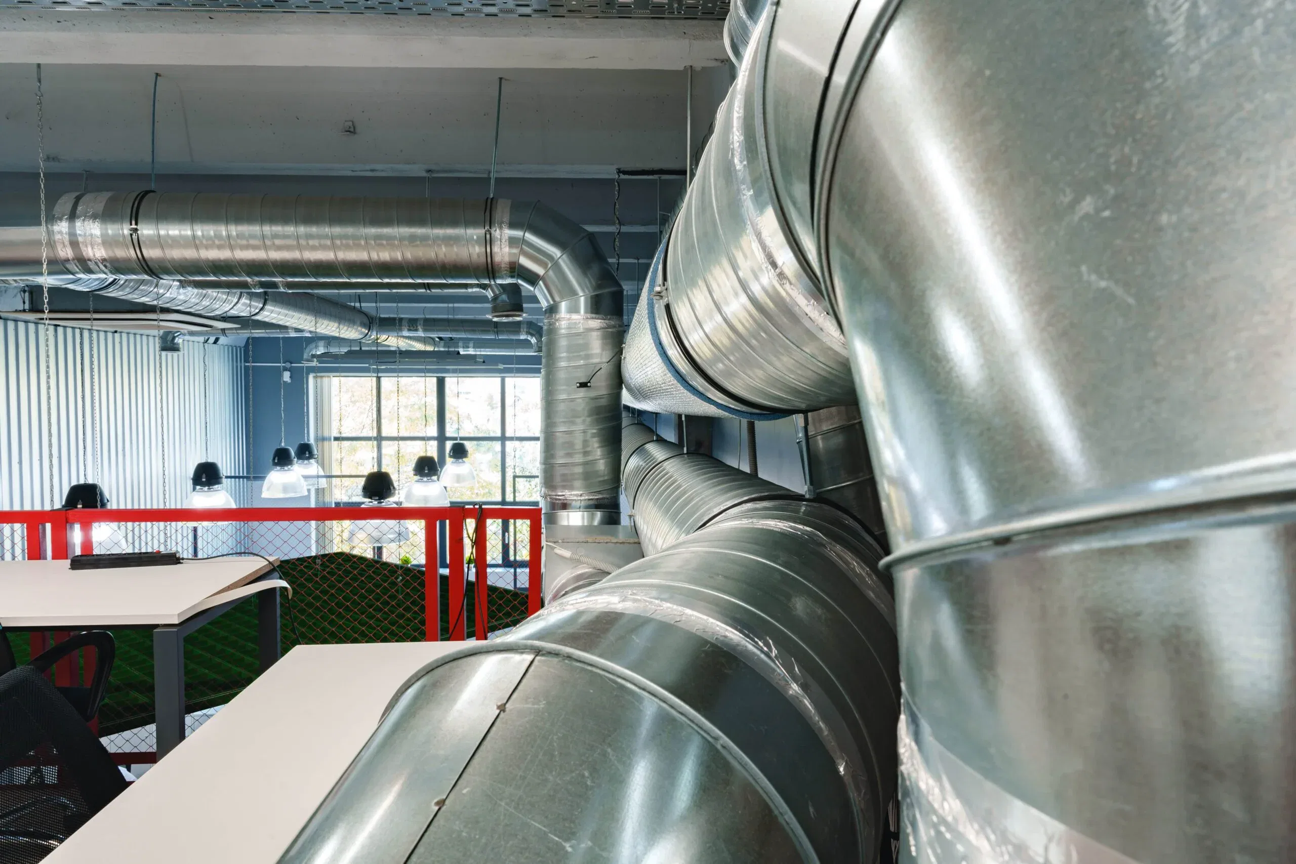 A perspective view of large, shiny silver galvanized steel ventilation ducts and spiral pipes hanging from the ceiling of an industrial or modern office space.