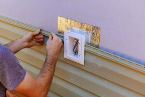 A close-up of a worker's hands precisely fitting a tan vinyl siding panel around an electrical outlet box on the exterior wall of a house during a renovation project.