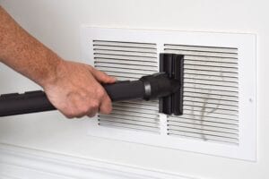 A person's hand using a black vacuum cleaner attachment to remove thick gray dust and lint from a white slatted return air vent on a wall.