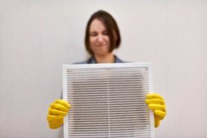 A woman in yellow gloves holding a dusty white ventilation grille during an indoor air quality inspection and cleaning service in Orlando, FL.