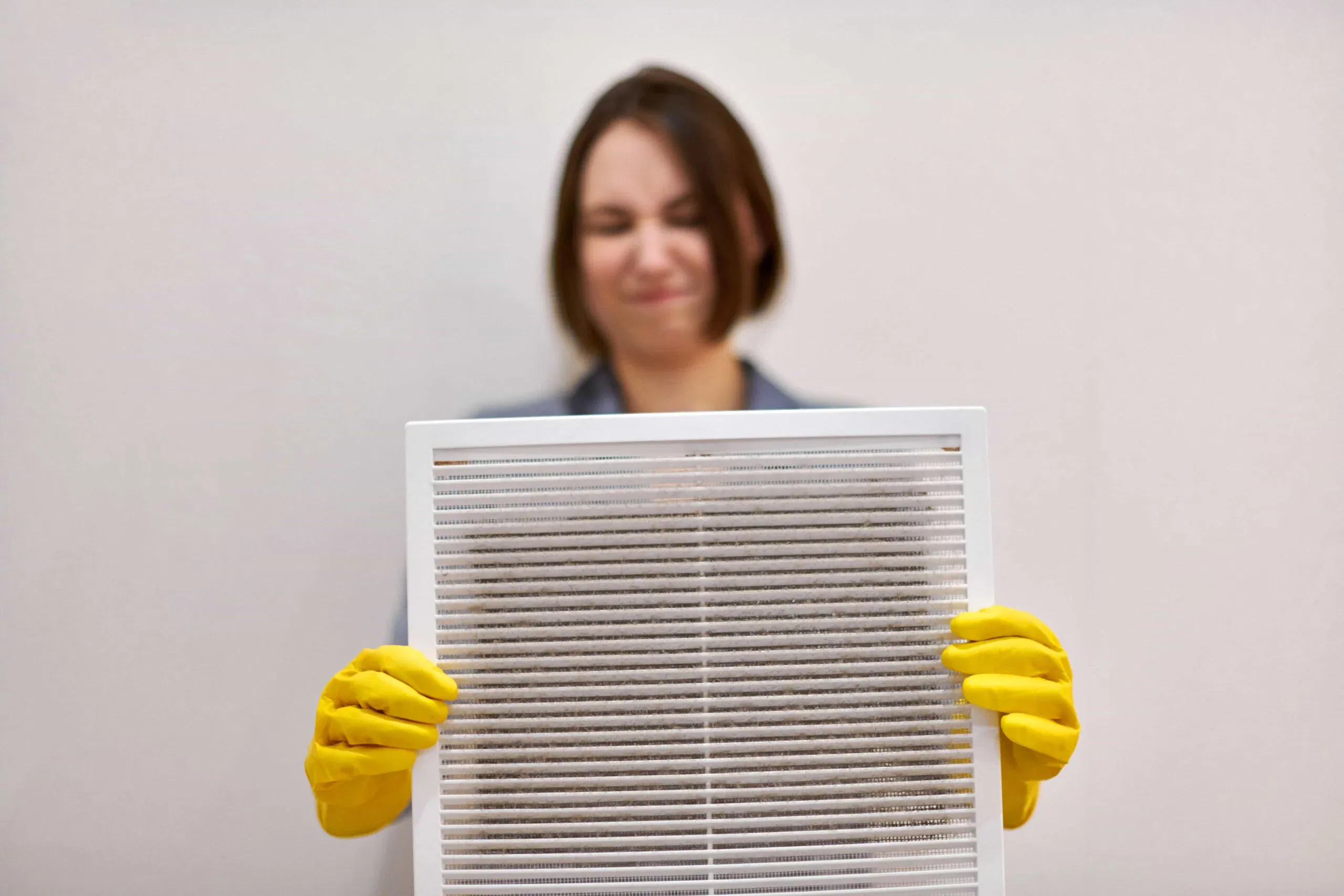 A woman in yellow gloves holding a dusty white ventilation grille during an indoor air quality inspection and cleaning service in Orlando, FL.