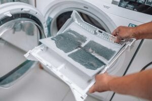 Close-up of a person's hands holding a white plastic dryer lint trap heavily covered in a thick layer of grey dust and fabric debris in front of an open dryer door.