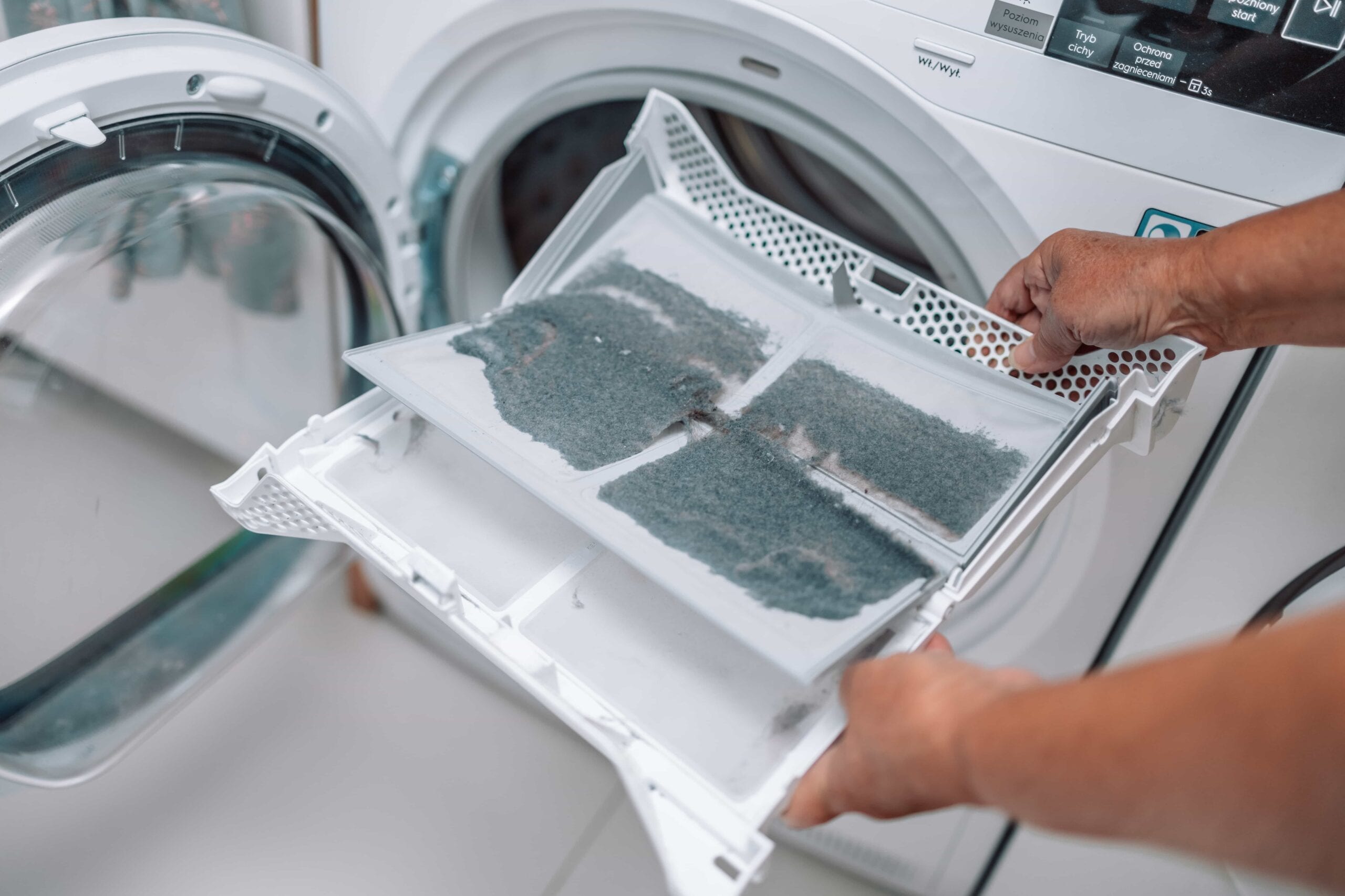 Close-up of a person's hands holding a white plastic dryer lint trap heavily covered in a thick layer of grey dust and fabric debris in front of an open dryer door.