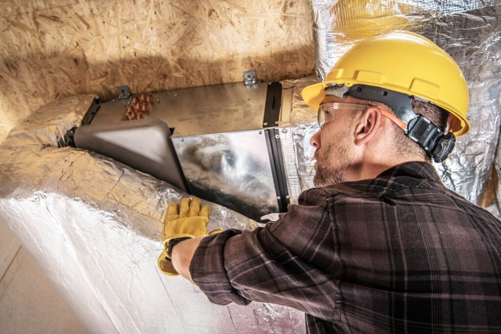 An HVAC worker in a yellow hard hat and safety glasses applying silver foil insulation wrap to metal air ducts in a residential attic.