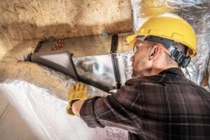 An HVAC worker in a yellow hard hat and safety glasses applying silver foil insulation wrap to metal air ducts in a residential attic.