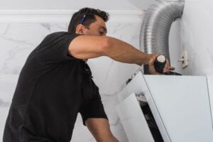A male contractor in a black shirt using a power drill and a spirit level to install a white modern kitchen cabinet against a marble-tiled wall near a silver ventilation duct.