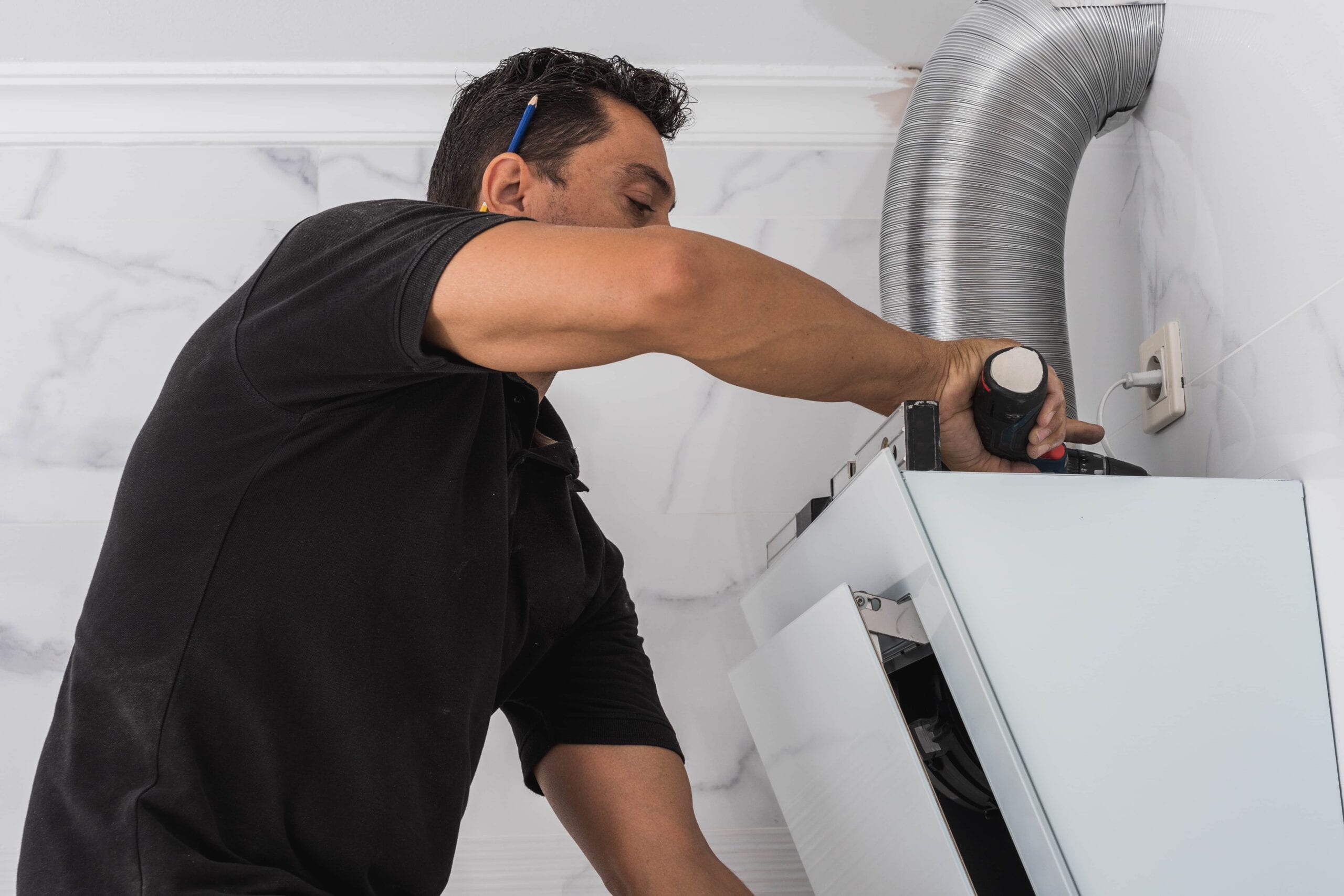 A male contractor in a black shirt using a power drill and a spirit level to install a white modern kitchen cabinet against a marble-tiled wall near a silver ventilation duct.