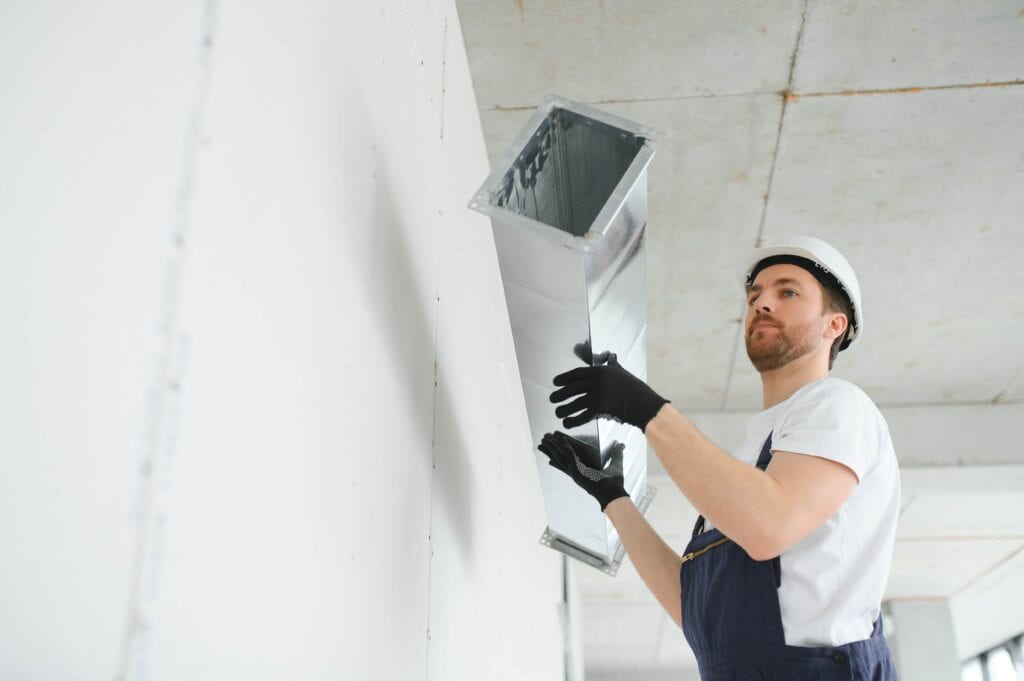 A professional HVAC technician wearing a white hard hat and gloves lifting a rectangular galvanized steel air duct into place during a commercial ventilation installation.