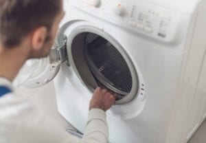 Over-the-shoulder view of a repair technician inspecting the rubber door seal and drum of a white front-load washing machine for mold or wear.