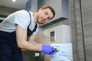 A professional repair technician in blue overalls and purple gloves servicing a front-loading washing machine in The Villages, FL.