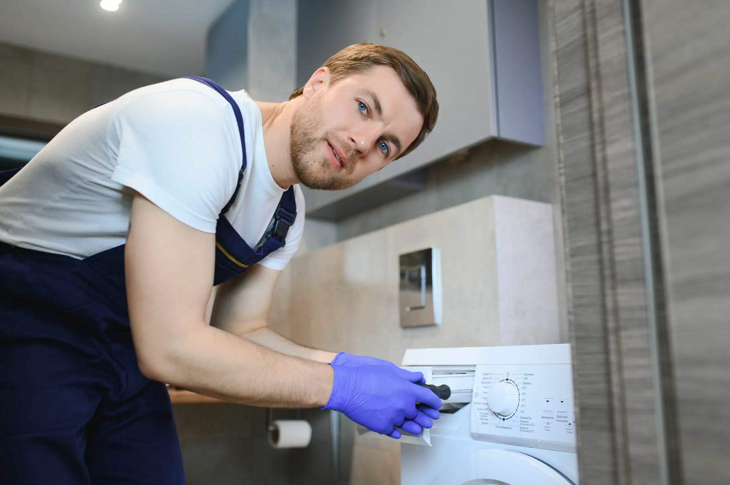 A professional repair technician in blue overalls and purple gloves servicing a front-loading washing machine in The Villages, FL.