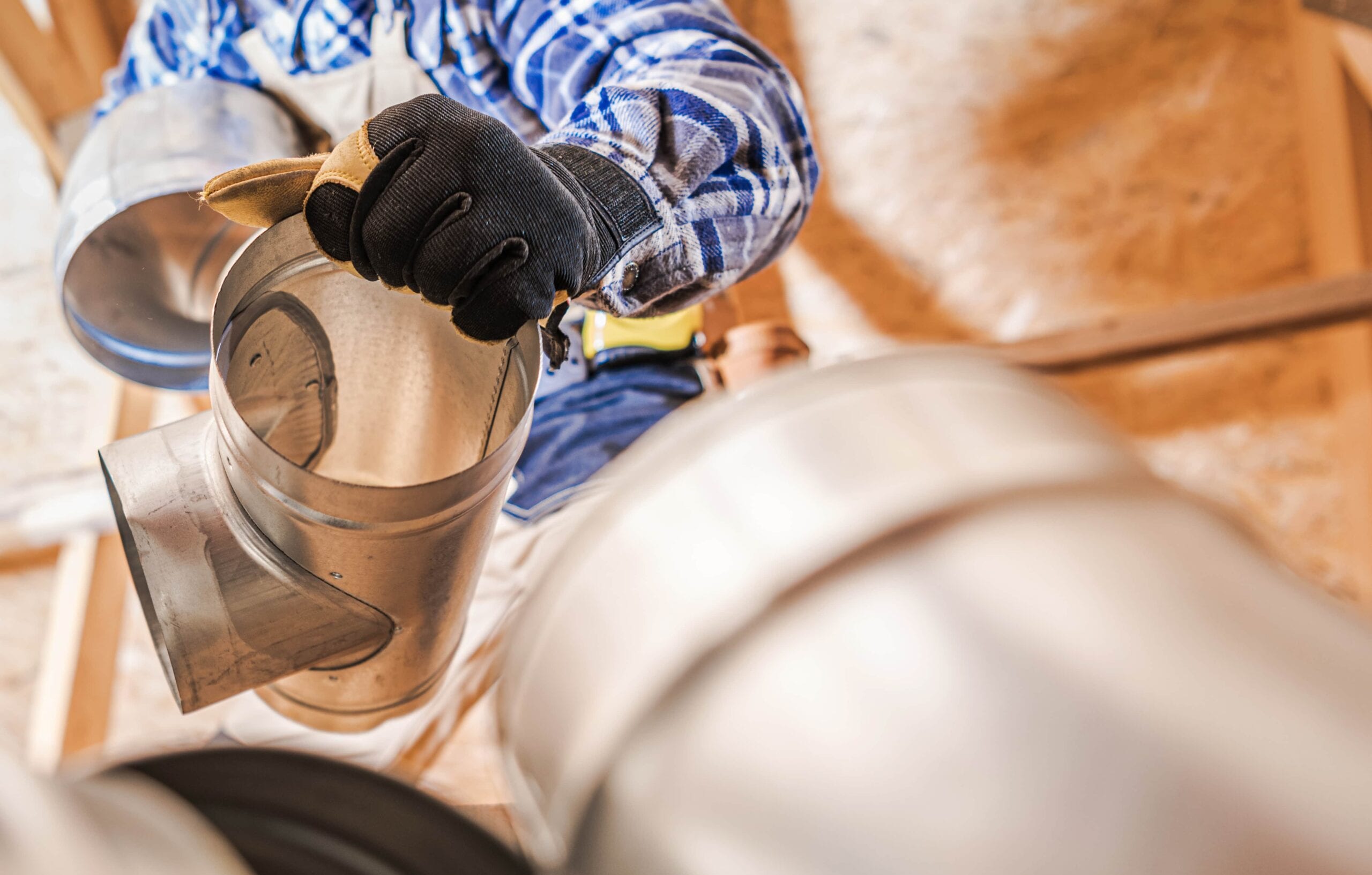 Close-up of a worker in a plaid shirt and work gloves assembling galvanized steel round ductwork and elbow joints in a residential attic space.