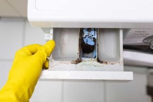 A gloved hand opening a dirty washing machine dispenser caked in mold and residue, highlighting critical laundry room maintenance and dryer vent cleaning in Florida.