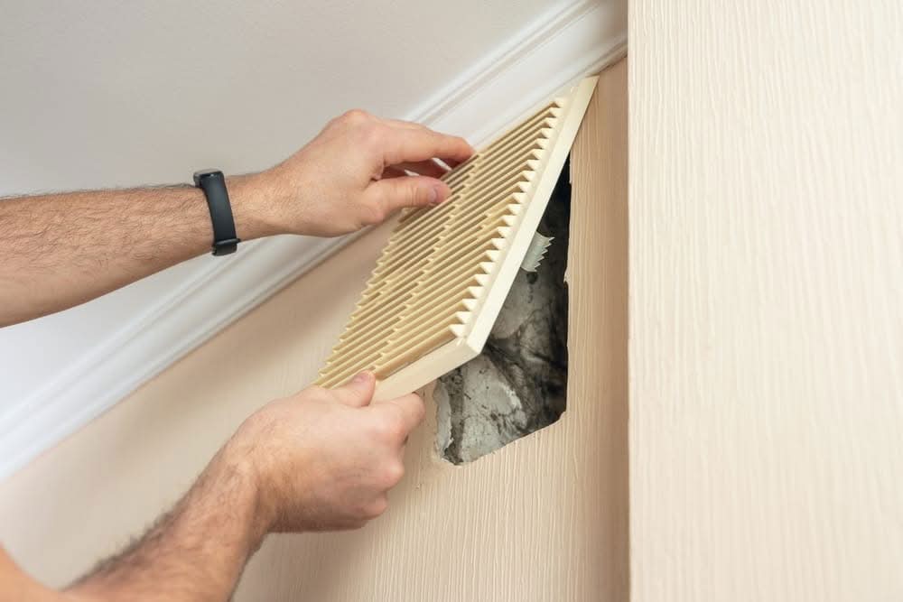 Close-up of a technician's hands removing an HVAC wall vent cover to reveal dusty ductwork during a home air quality inspection in Lakeland, FL.